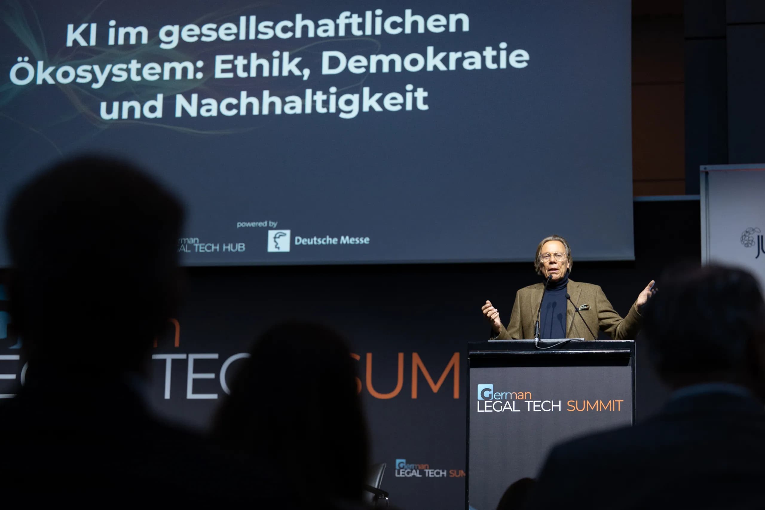 Blick aus dem Publikum auf einen Redner am Pult, darüber die Leinwand „KI im gesellschaftlichen Ökosystem“